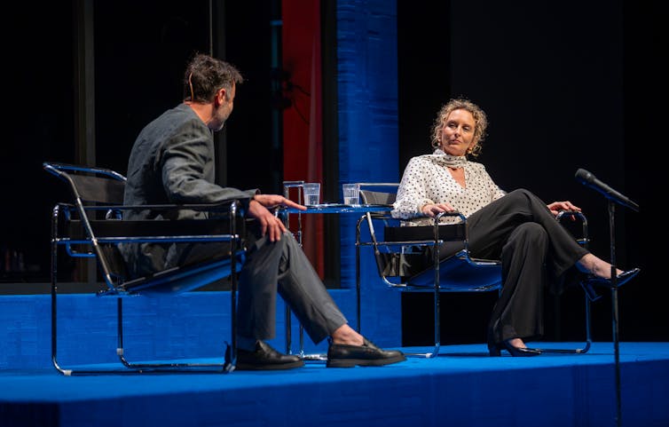 Production image: a man and a woman talk on a panel.