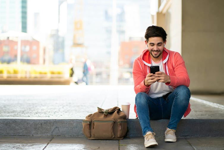 A young man in jeans and a red sweater sits on a step texting on his phone and smiling.