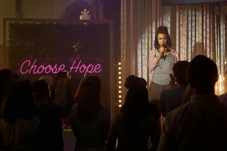 woman stands on stage wiht microphone, dark lighting, bright pink wording in lights say 'choose hope'