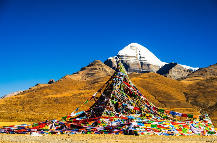 A snow-capped mountain rises among a range of peaks, with Buddhist prayer flags lining the path leading toward it.