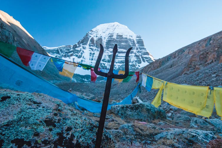 Colourful prayer flags and a trident, symbolizing the Hindu god Shiva, placed in front of a snow-capped mountain.