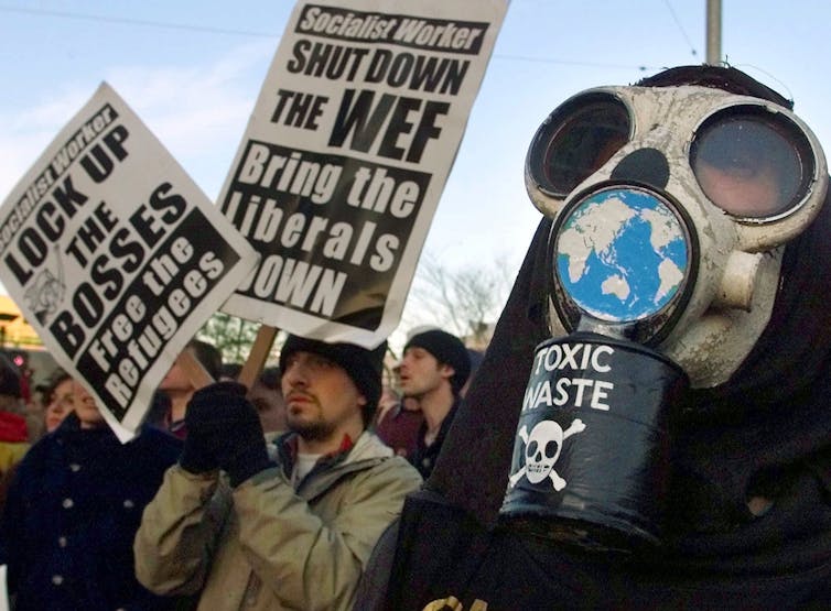 A person wearing a gas mask and another holding two signs reading 'shut down the WEF' and 'Lock up the bosses'