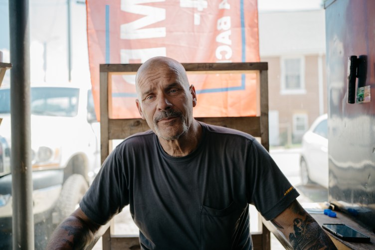 A bald man wearing a gray T-shirt and with tattoos on his forearms stares directly at the camera. Behind him a wooden pallet leans against a plate-glass window, with a Trump 2024 flag hanging outside.
