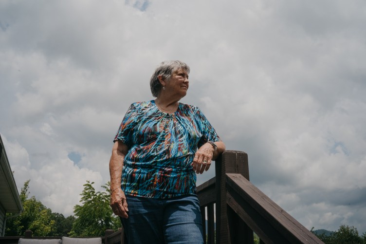 A woman with short gray hair stares off into the distance. She is standing on a deck outside as white clouds cover the sky.