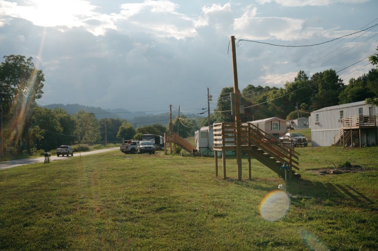 A wooden staircase unconnected to any structure stands on a green lawn next to a utility pole. Trailers and cars can be seen behind it.