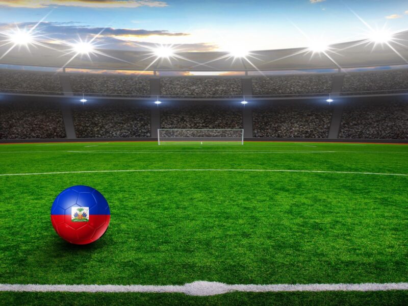 Feature image of a football in the colours of the Haitian flag, on a soccer field, created using Canva Pro elements. 