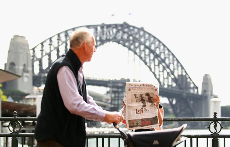 A grey-haired man pushes a pram along a path in front of the Sydney Harbour Bridge, while a woman in the background reads a newspaper.