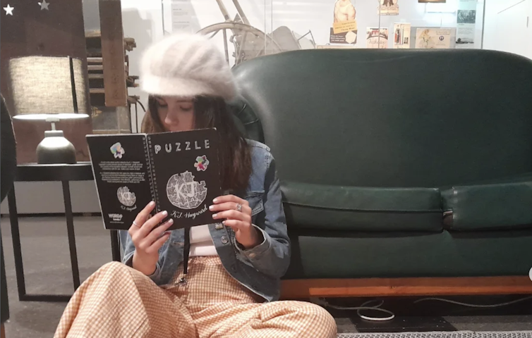 A young woman reading a book in front of a lounge, wearing a white peaked cap.