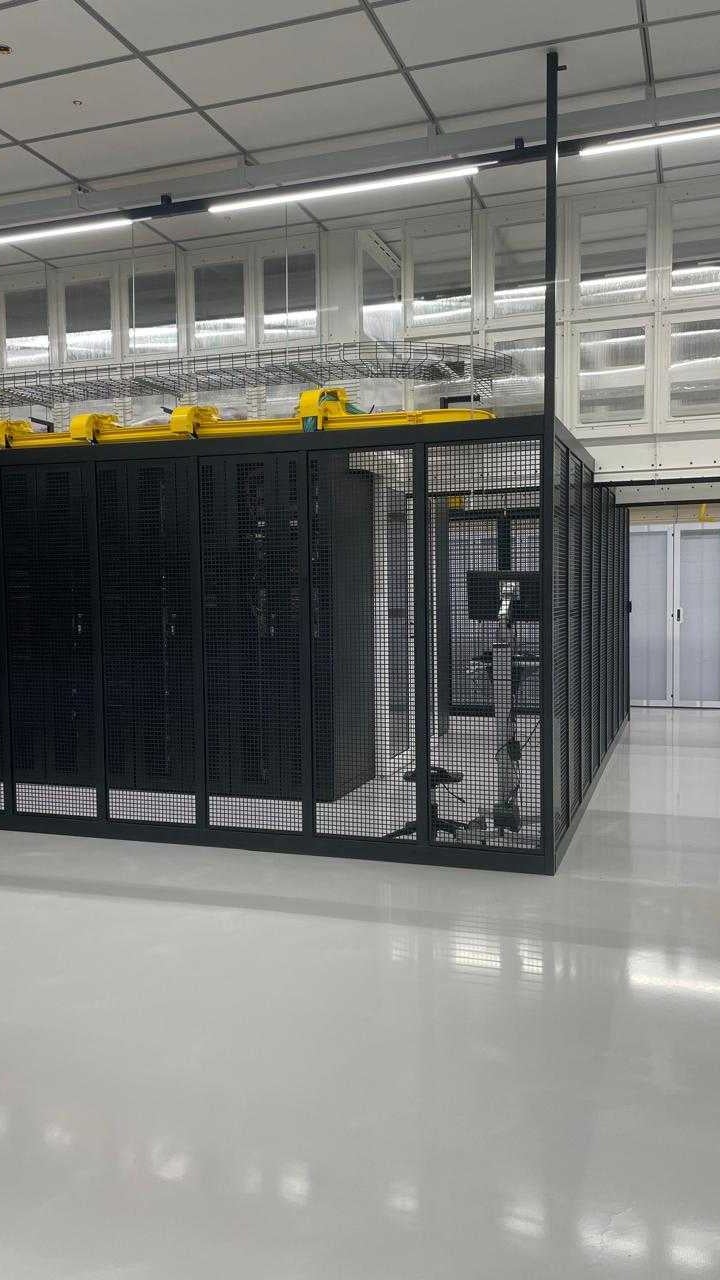 A spacious server room featuring rows of black mesh server racks, bright lighting, and a clean white floor
