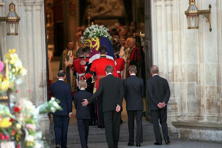 Three men and two boys walk behind a coffin in a funeral procession, one man with a hand on a boy's back to comfort him.