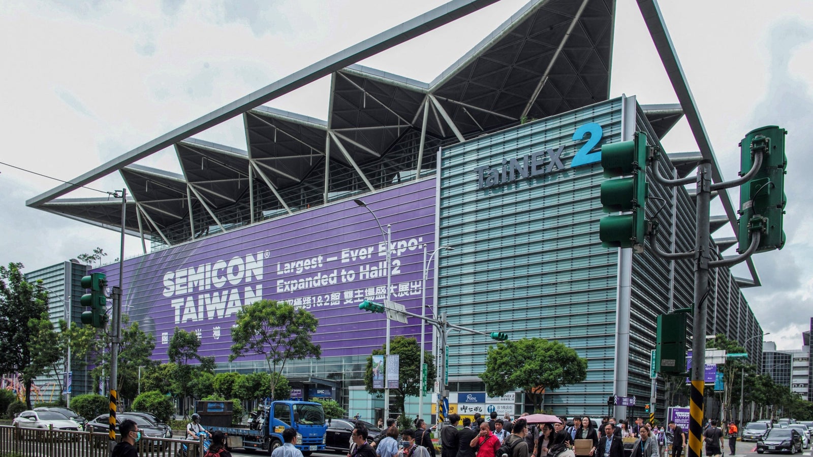 A busy street in front of the TaiNEX 2 exhibition center with a large banner for SEMICON TAIWAN, featuring pedestrians and traffic signals