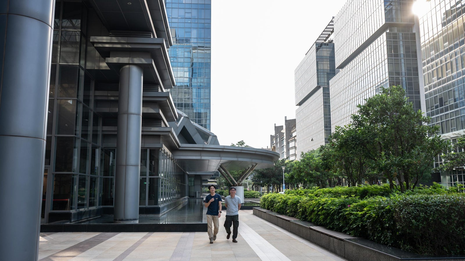 Two men casually walk along a landscaped pathway near modern glass buildings in an urban setting