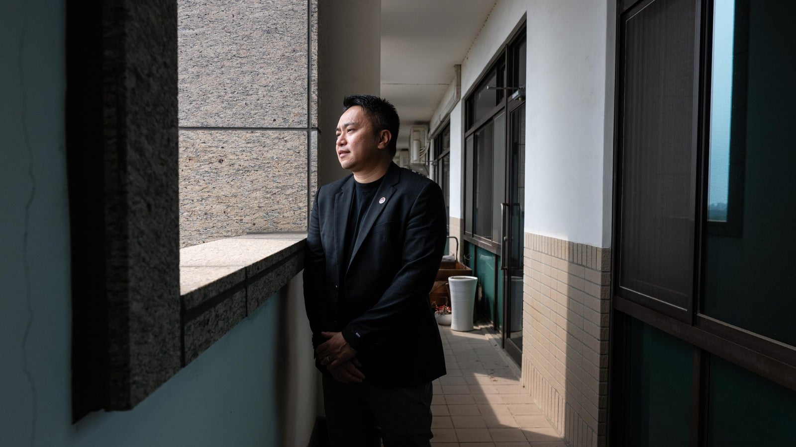 A man in a black blazer stands by a window, gazing thoughtfully outside, with a corridor and potted plants in the background