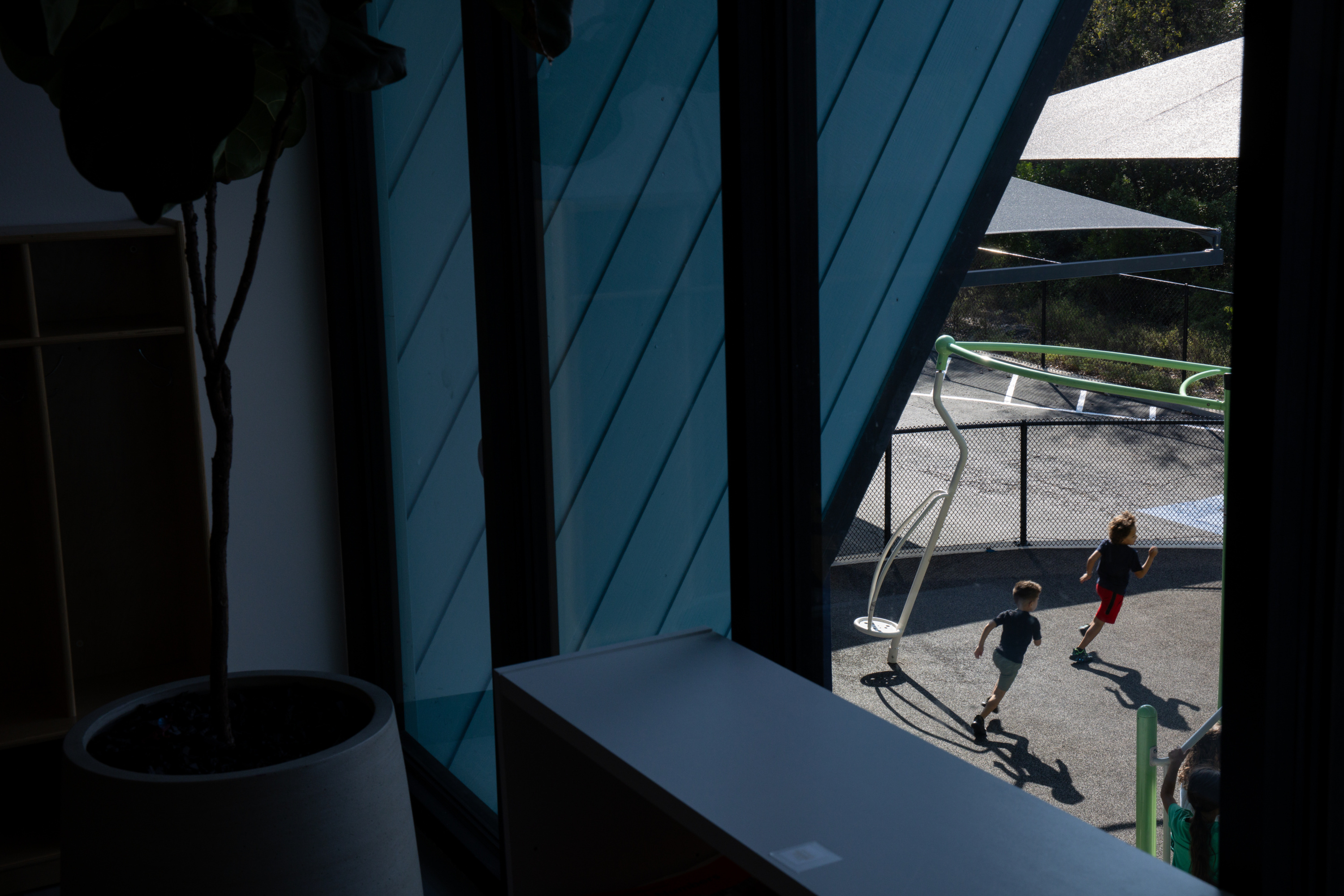 Two small children can be seen through a window running near playground equipment. The interior of the room is blue, and a potted plant sits in front of the window.