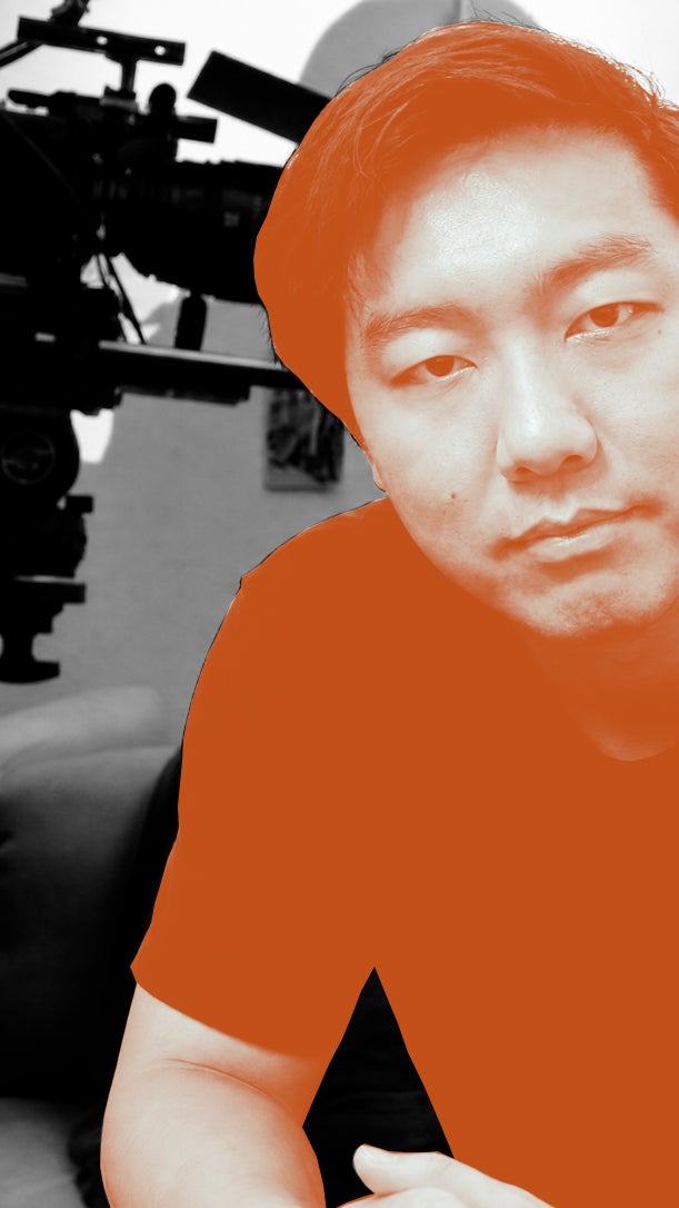 A person with short hair is seated in front of a film camera, portrayed in high contrast with an orange overlay
