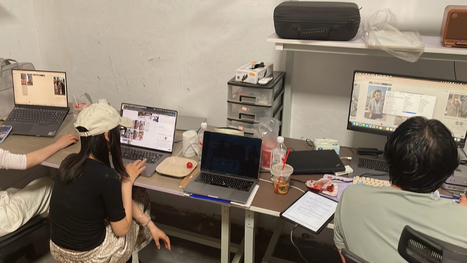 A group of individuals working on laptops at a shared workspace, with various personal items and drinks on the table