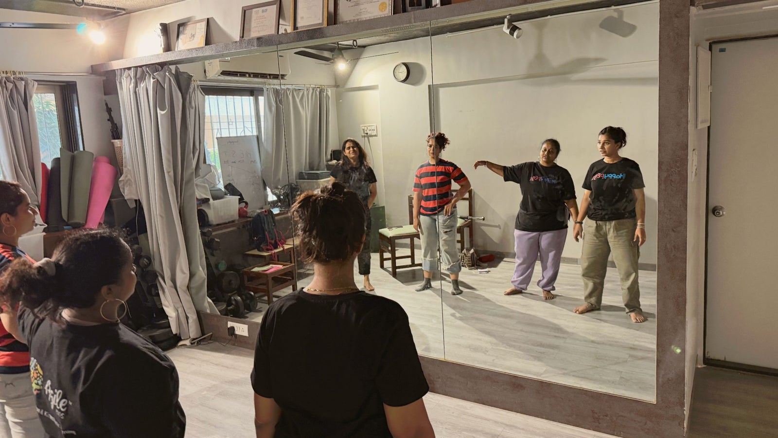 A group of five women is practicing in a dance studio, facing a mirror, with one woman demonstrating a movement