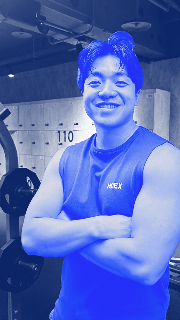 Smiling individual with crossed arms stands in a gym, showcasing a muscular build. Exercise equipment and lockers are visible