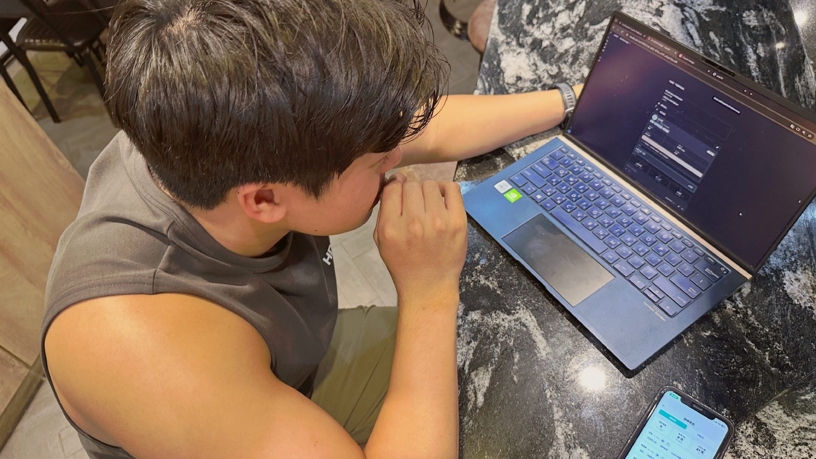 A person seated at a table, focused on their laptop, with a smartphone displaying information nearby