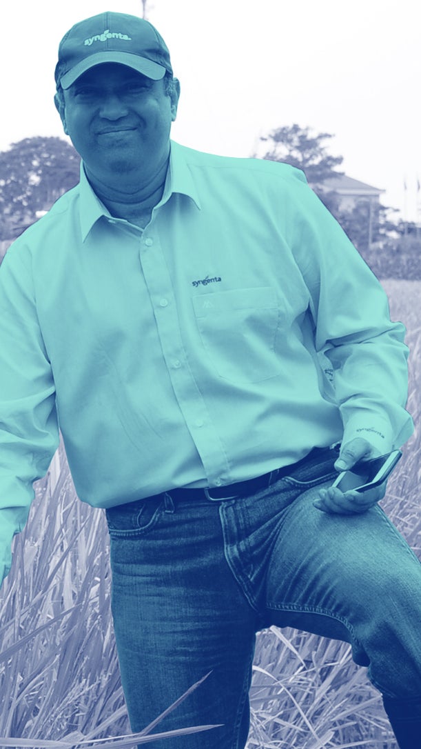 A man in a light blue shirt and cap stands in a rice field, smiling while holding a phone and examining a plant