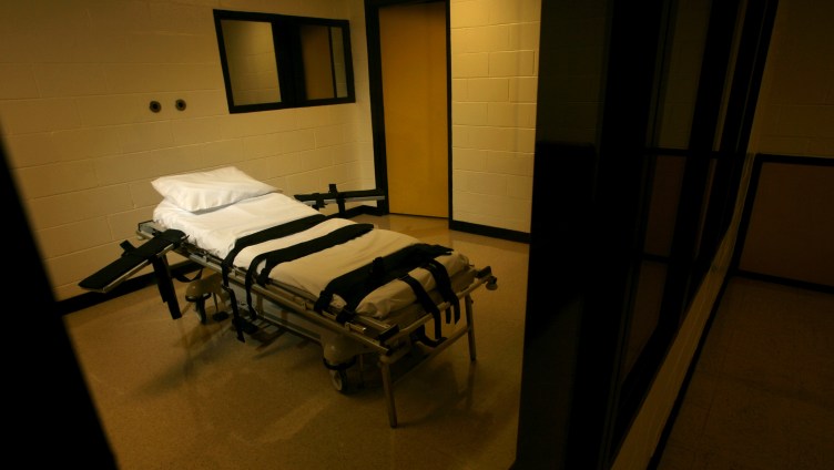 A gurney with black straps, a white cot and white pillow sits in an empty, yellow, dimly lit room.