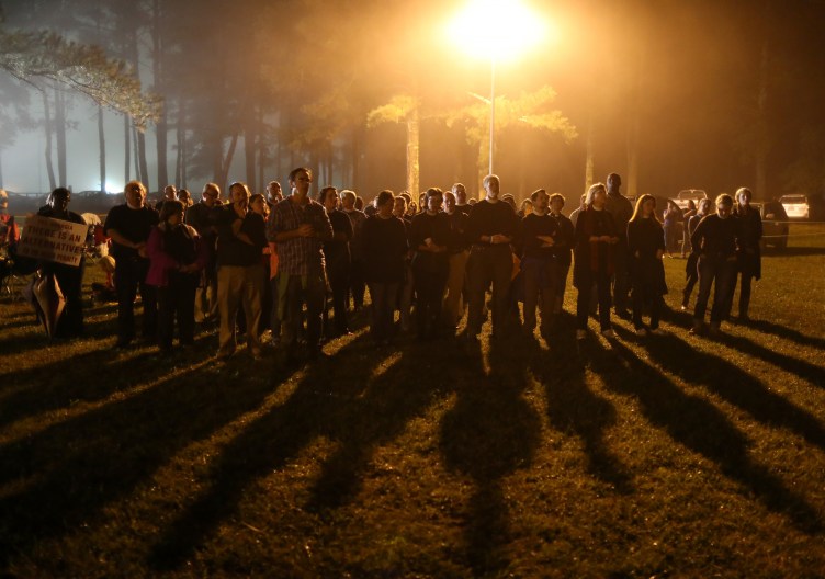 A crowd of people stand in front of shadowy woods and a yellow street light, casting elongated shadows onto the lawn in front of them.