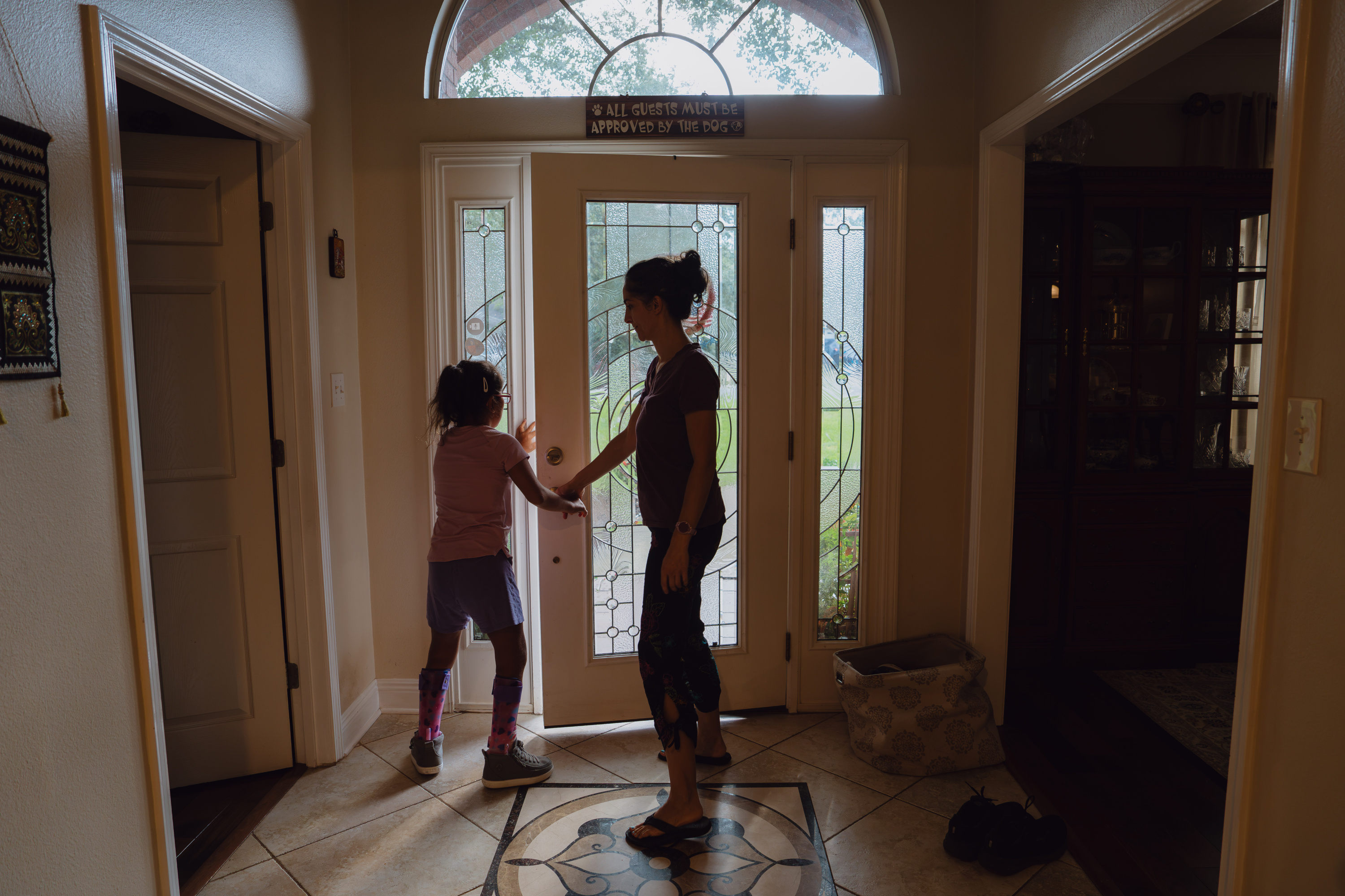 A woman and a child are silhouetted in front of a glass front door in a large tiled entryway. A sign above the door reads, “All guests must be approved by the dog.”