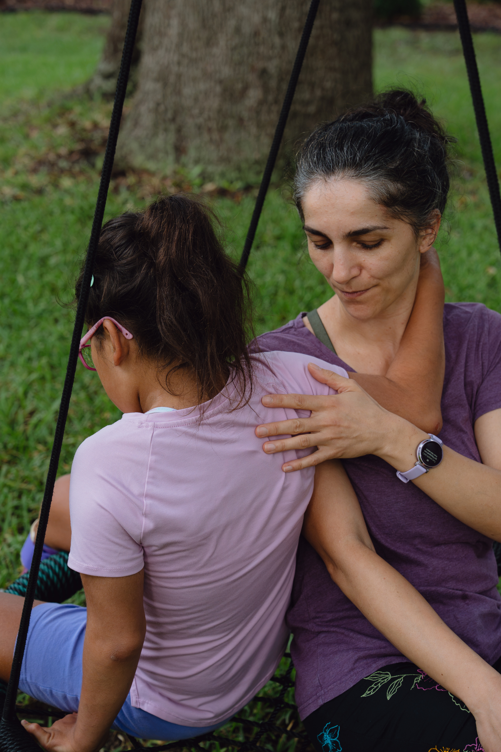 A woman wearing a purple shirt and ponytail puts her arm around Amelia, who is facing away from the camera. They are sitting on a rope swing outside.