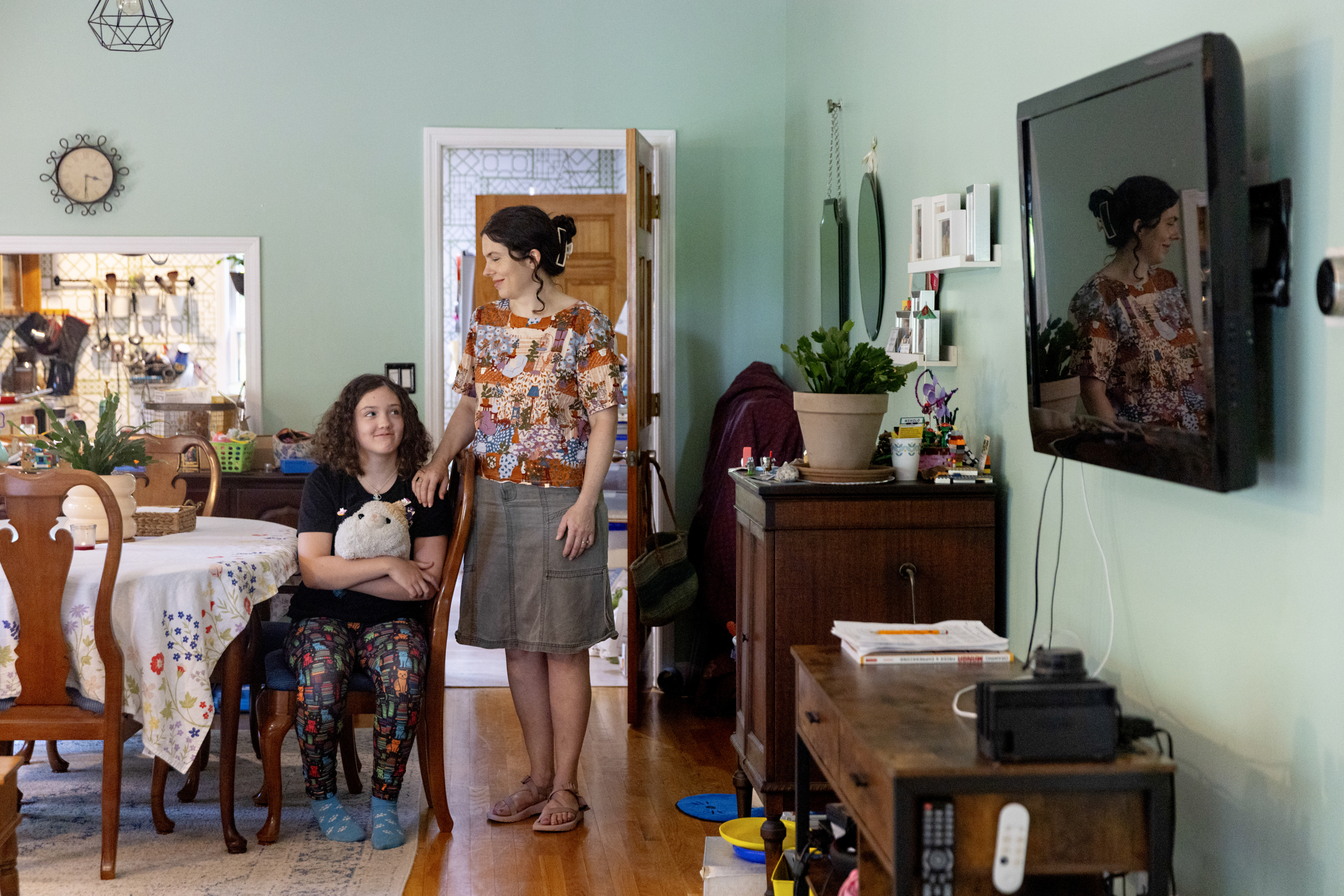 A girl wearing a black shirt and leggings decorated with cats sits at a dining room table with a cat stuffed animal on her lap. A woman wearing a patchwork blouse stands next to her and holds her shoulder.