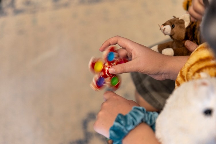 Hands play with a fidget spinner. A blue scrunchie is on one wrist. Stuffed animals appear in the frame.