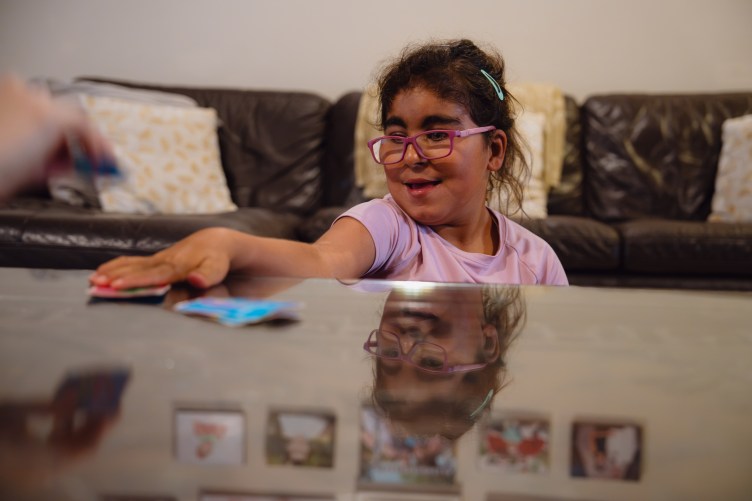 A smiling girl reaches her hand over a stack of cards on a shiny brown coffee table. Her reflection can be seen in the table’s surface.