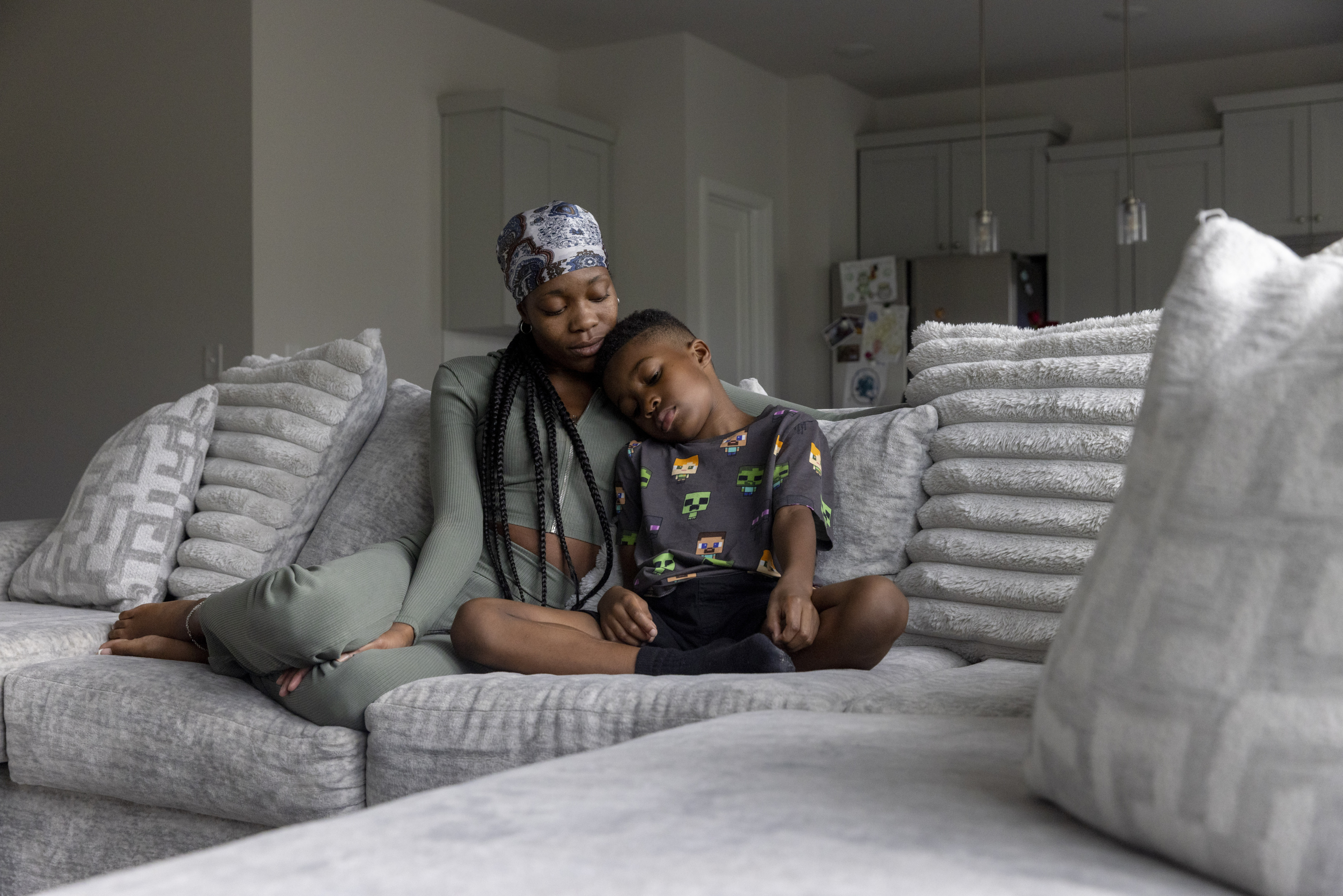 A woman wearing a headscarf and a sage green outfit sits on a gray couch with many pillows next to a boy wearing a black shirt with a colorful pattern.