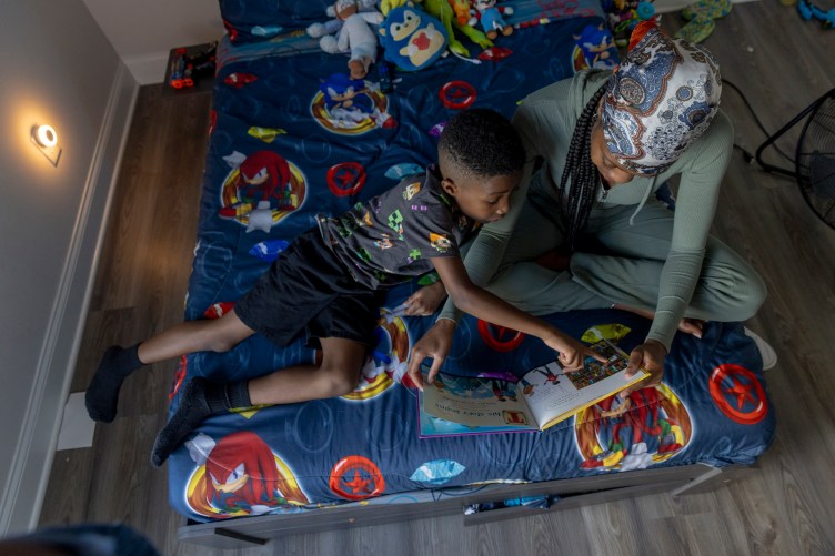 Overhead view of a woman sitting crosslegged, holding a book open as a boy sits next her and points at the book. The bedspread has characters from Sonic games, and stuffed animals sit in the background.