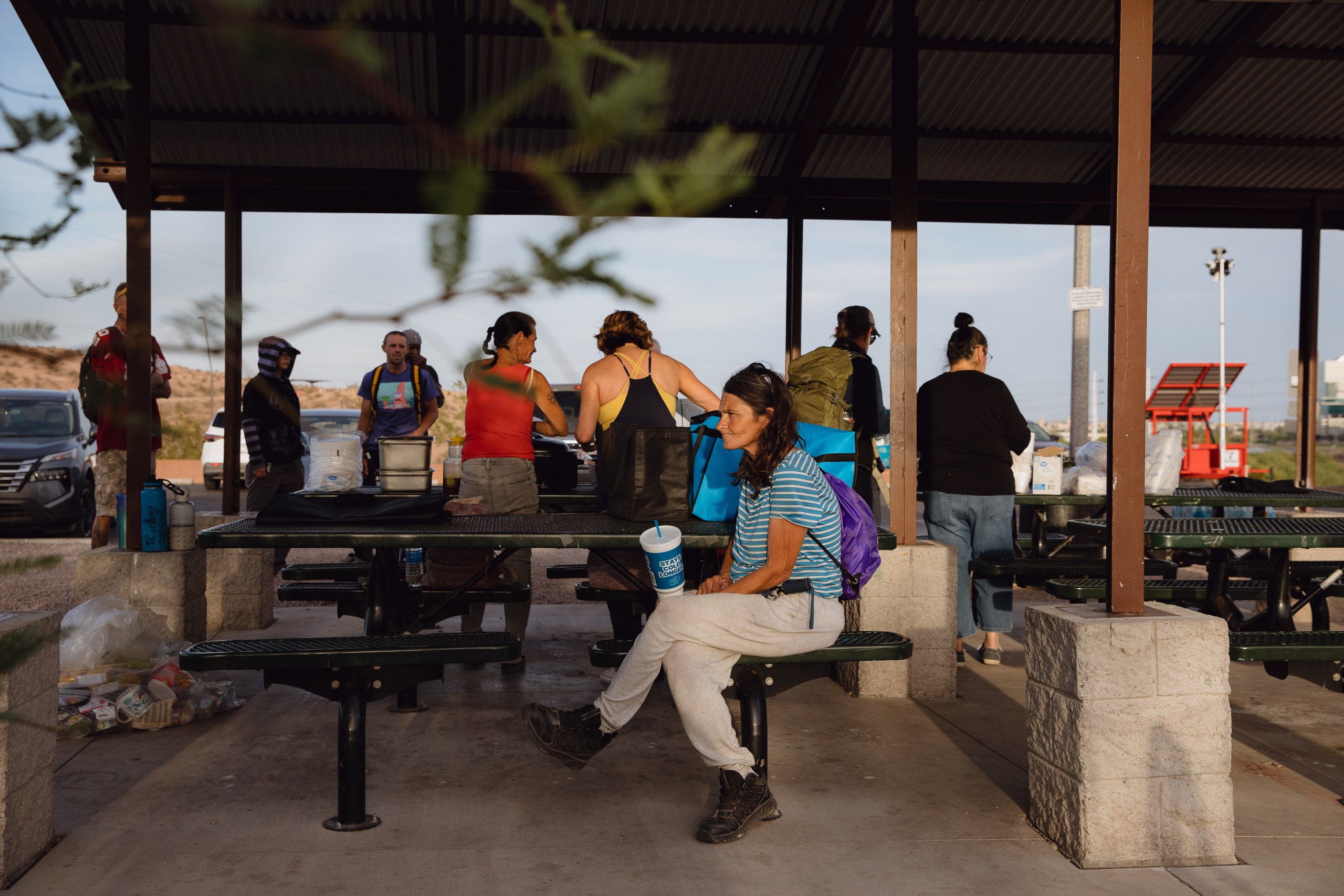 A woman wearing a backpack sits at a picnic table under a gazebo while several men and women wait in a line to be served food by two volunteers.