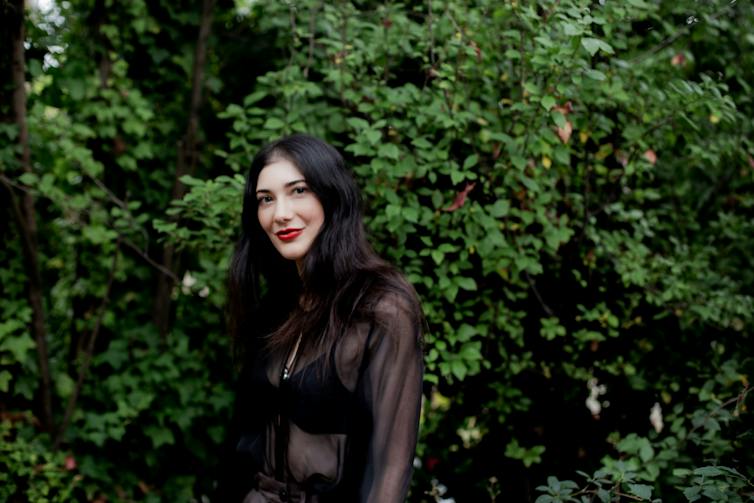 The author, Laura Elizabeth Woollett, a beautiful woman with long, dark hair and red lipstick in front of a hedge.