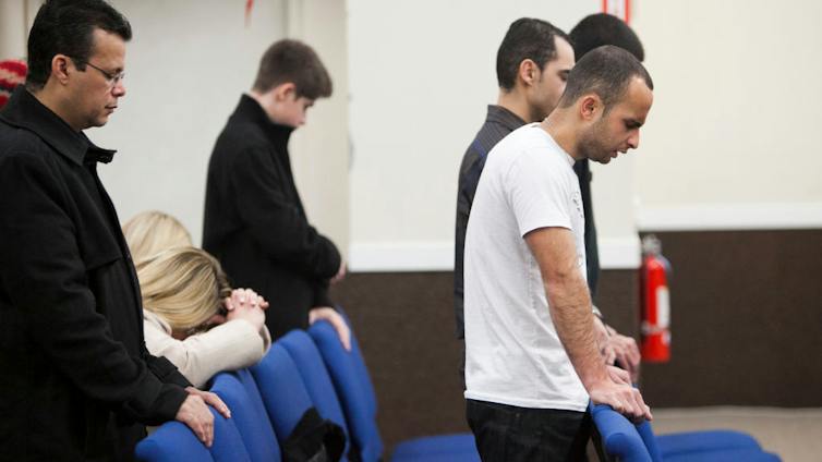 Several men bow their heads in prayer as they stand behind blue chairs.