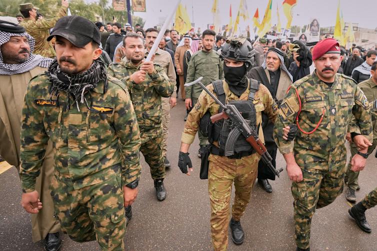 Iraqis in military uniforms, one wearing a balaclava, walk along a Baghdad street.