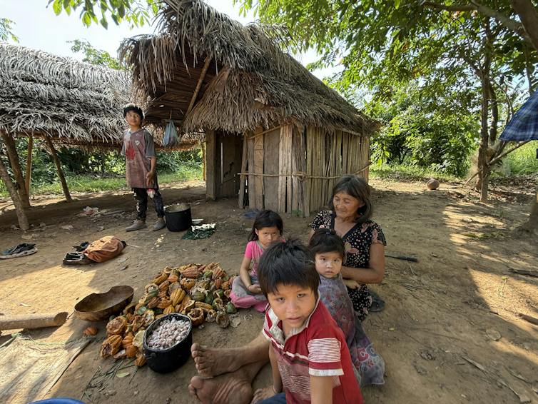 several children sit with a woman next to some harvested crops in front of a thatched hut