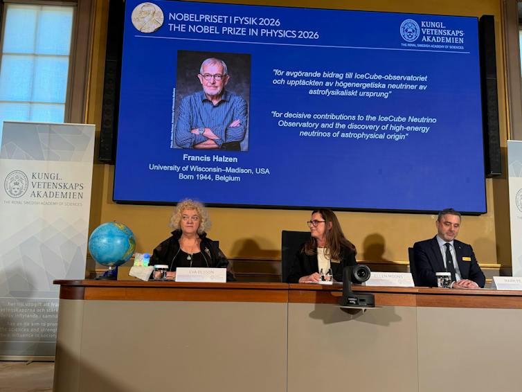 A panel of three people sitting at a table, with a screen behind them showing a photo of Francis Halzen and a description of his research.