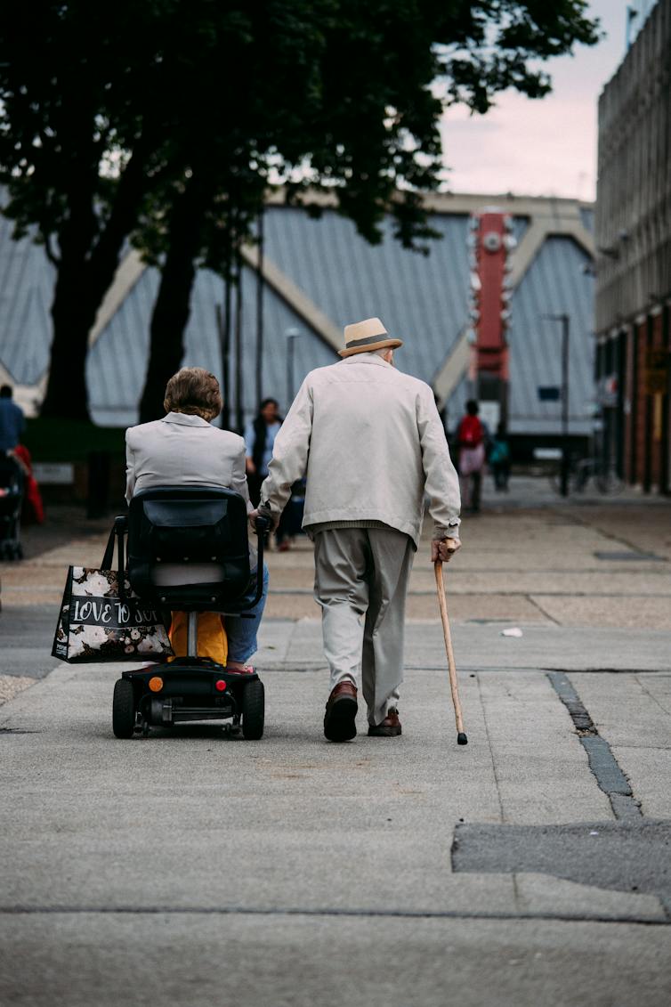 A man walks with a cane while a woman moves alongside him on a mobility scooter