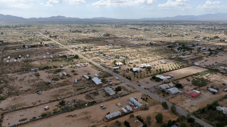 An aerial view captures a sprawling desert community with roads cutting through sparse terrain, scattered homes and mobile residences leading toward distant mountain ranges.