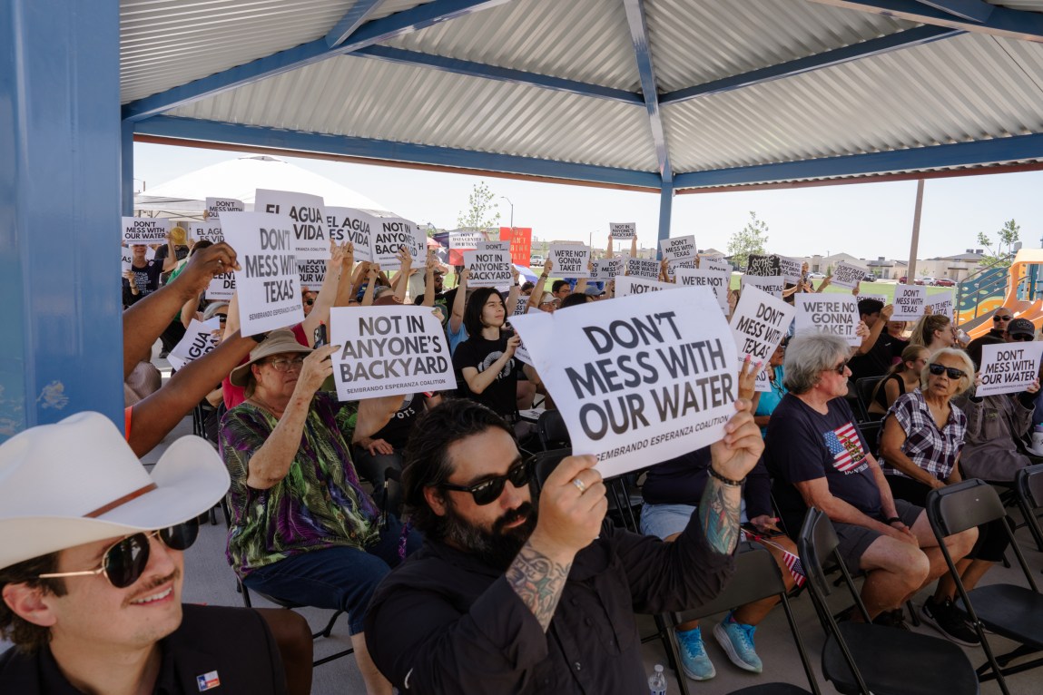 A crowd gathered under a covered outdoor pavilion holds up printed signs with slogans like, "Don’t mess with our water," "Not in anyone’s backyard," and “El agua es vida.”