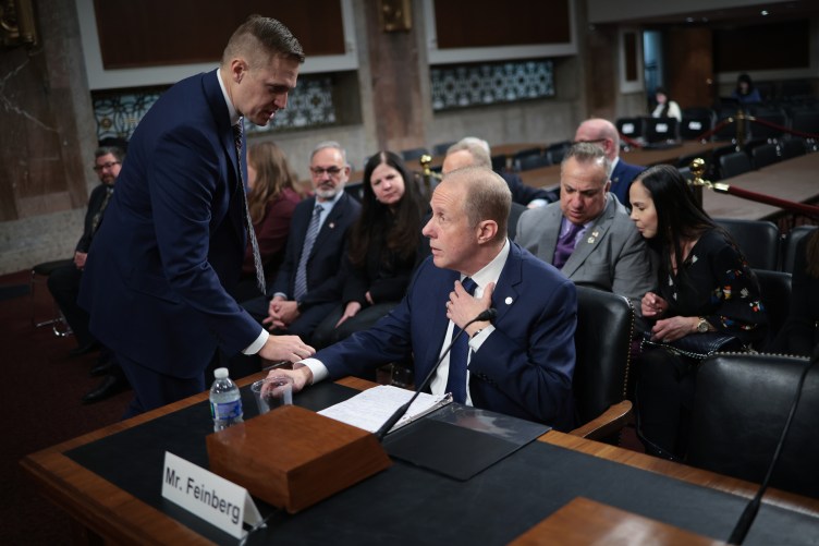 A seated man in a suit confers with a man who is standing and leaning in to talk to him. A row of people are seated behind them in a government building.